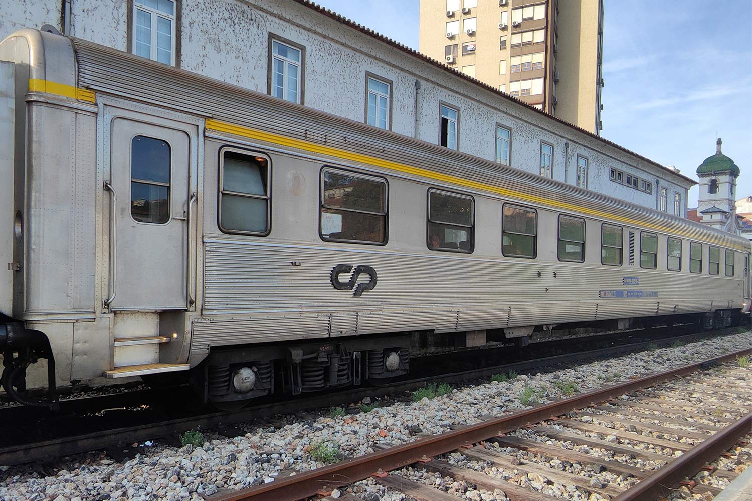 Carruagem Sorefame de 1ª classe cor cinzento e com lista amarela por cima das janelas, parqueada numa das linhas da estação.