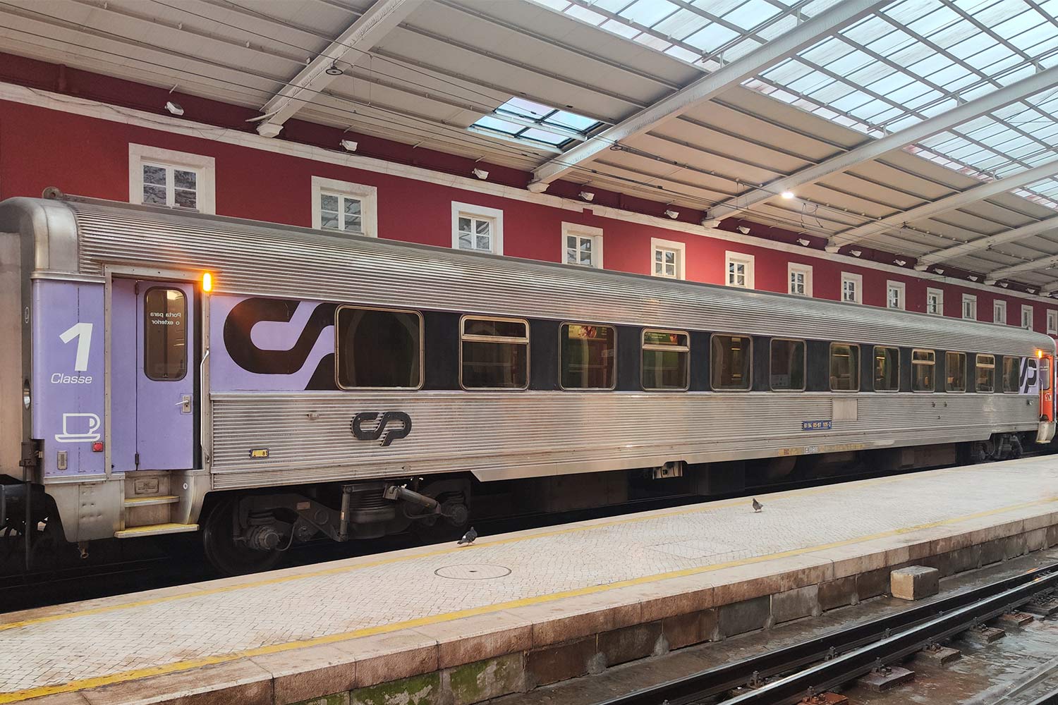 Carruagem Sorefame de 1ª classe com bar cor roxo e cinzento parqueada numa das linhas da estação.