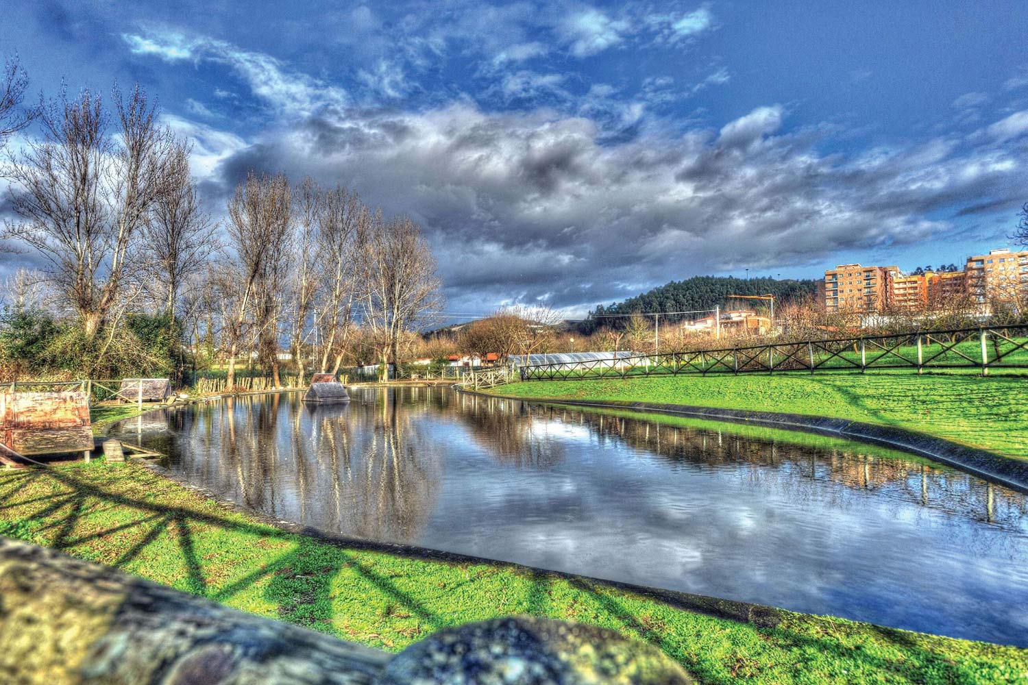 Imagem da quinta com lago, relva e árvores sem folhas, céu nublado com fundo de prédios e floresta