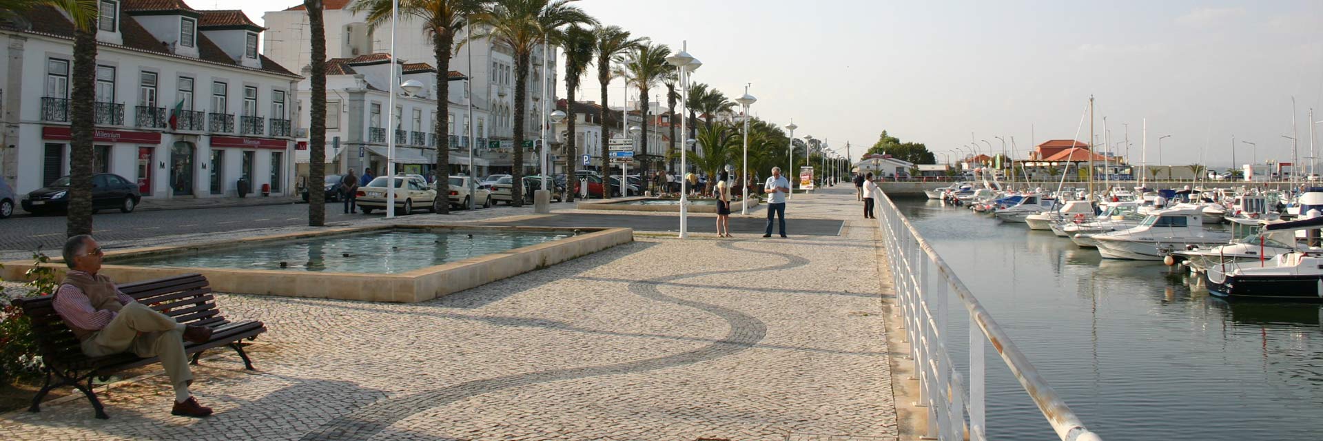 Zona ribeirinha de Vila Real de Santo António, com barcos atracados e pessoas sentadas e a caminhar pelo passeio com palmeiras.