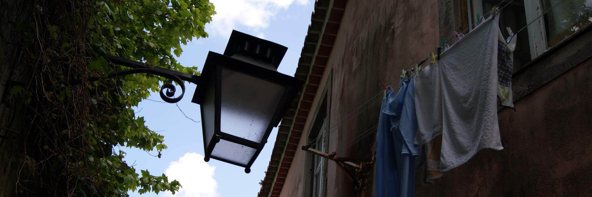 Pormenor de um candeeiro de rua em grande plano, entre parede com arvoredo e prédio com roupa estendida na janela