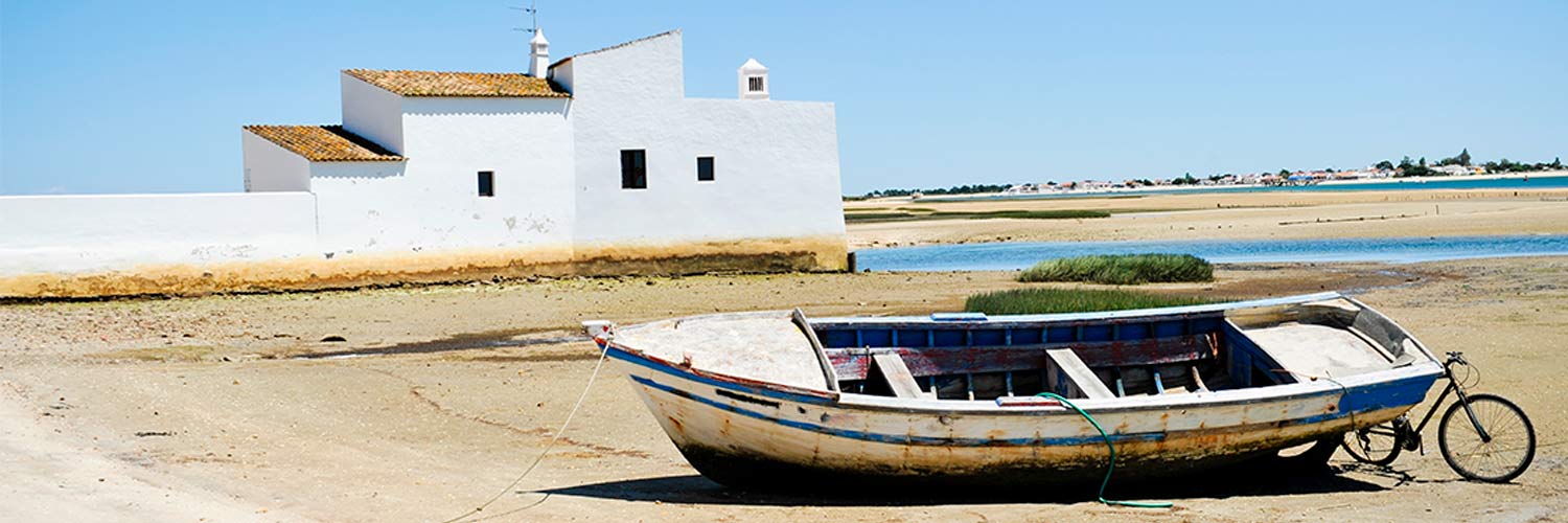 a boat with a bicycle leaning against it and a white building on the sandy shore of the Ria Formosa