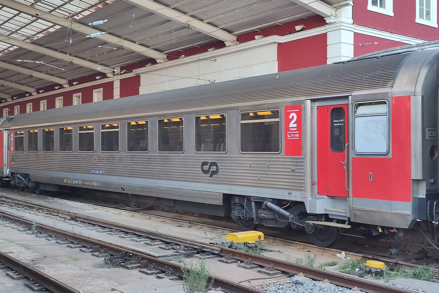 Carruagem Corail de 2ª classe cor vermelho e cinzento parada numa das linhas da estação.