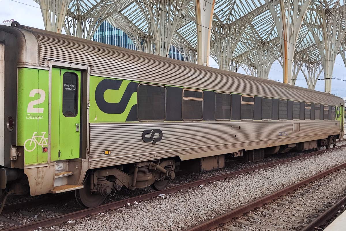 Carruagem Sorefame de 2ª classe cor verde e cinzento parqueada numa das linhas da estação.