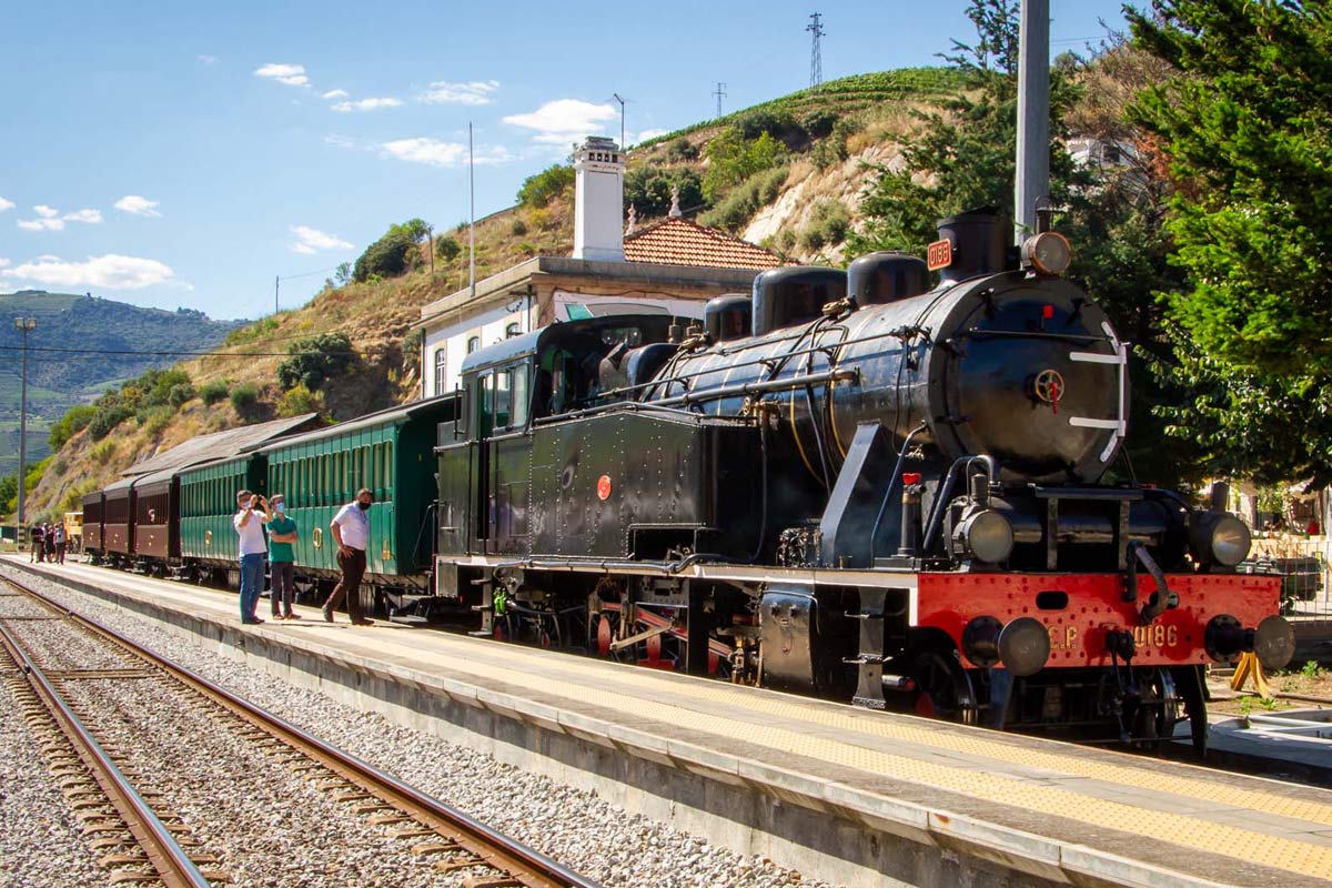 Comboio Histórico do Douro, formado por locomotiva a vapor CP 0186, cor preto e vermelho, e cinco carruagens de madeira, cor castanho e verde, parado na estação.