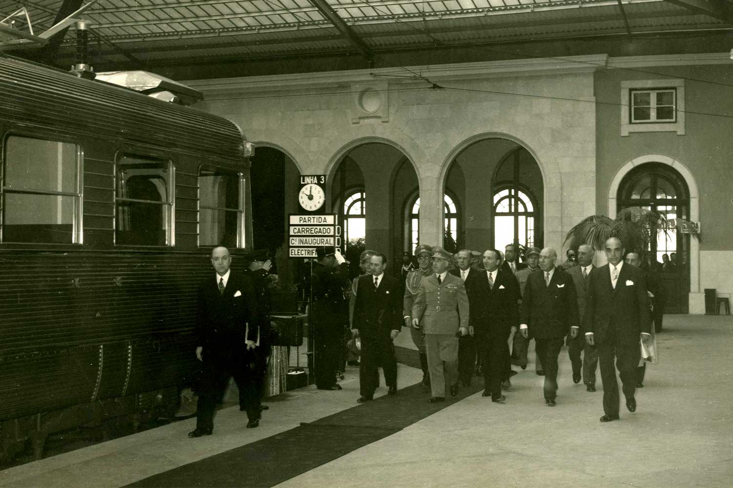 Imagem a preto e branco da estação de Santa Apolónia, com pessoas a caminhar junto a comboio elétrico e perto da plataforma de embarque.