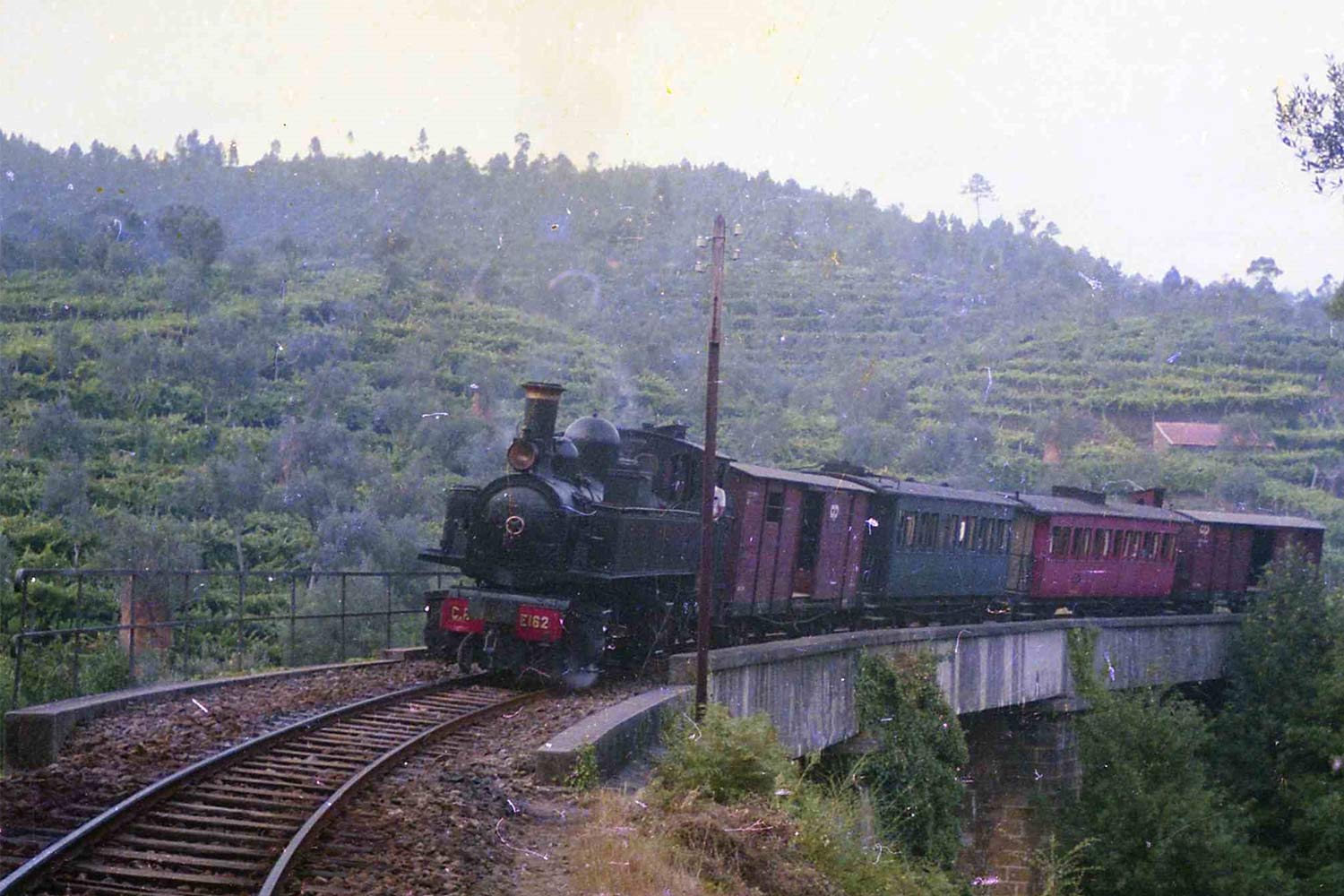 Locomotiva a vapor de via métrica CP E162, cor preto, a rebocar comboio regional formado por duas carruagens de passageiros cor vermelho e azul, e dois vagões de mercadorias, cor castanho.