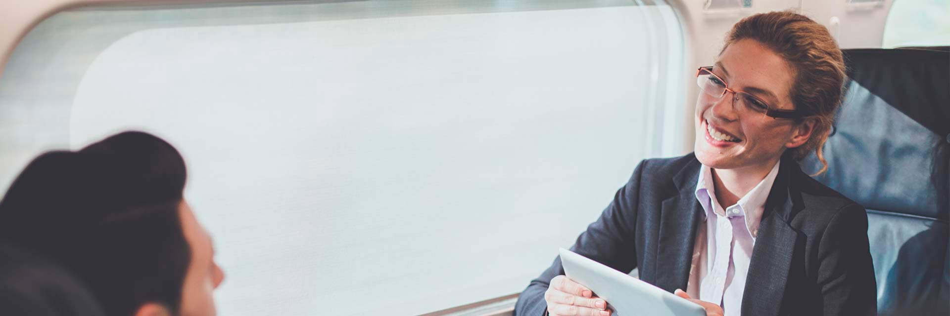 Mulher executiva com tablete e homem ao telemóvel, frente a frente numa mesa de reuniões, junto à janela, no Alfa Pendular.