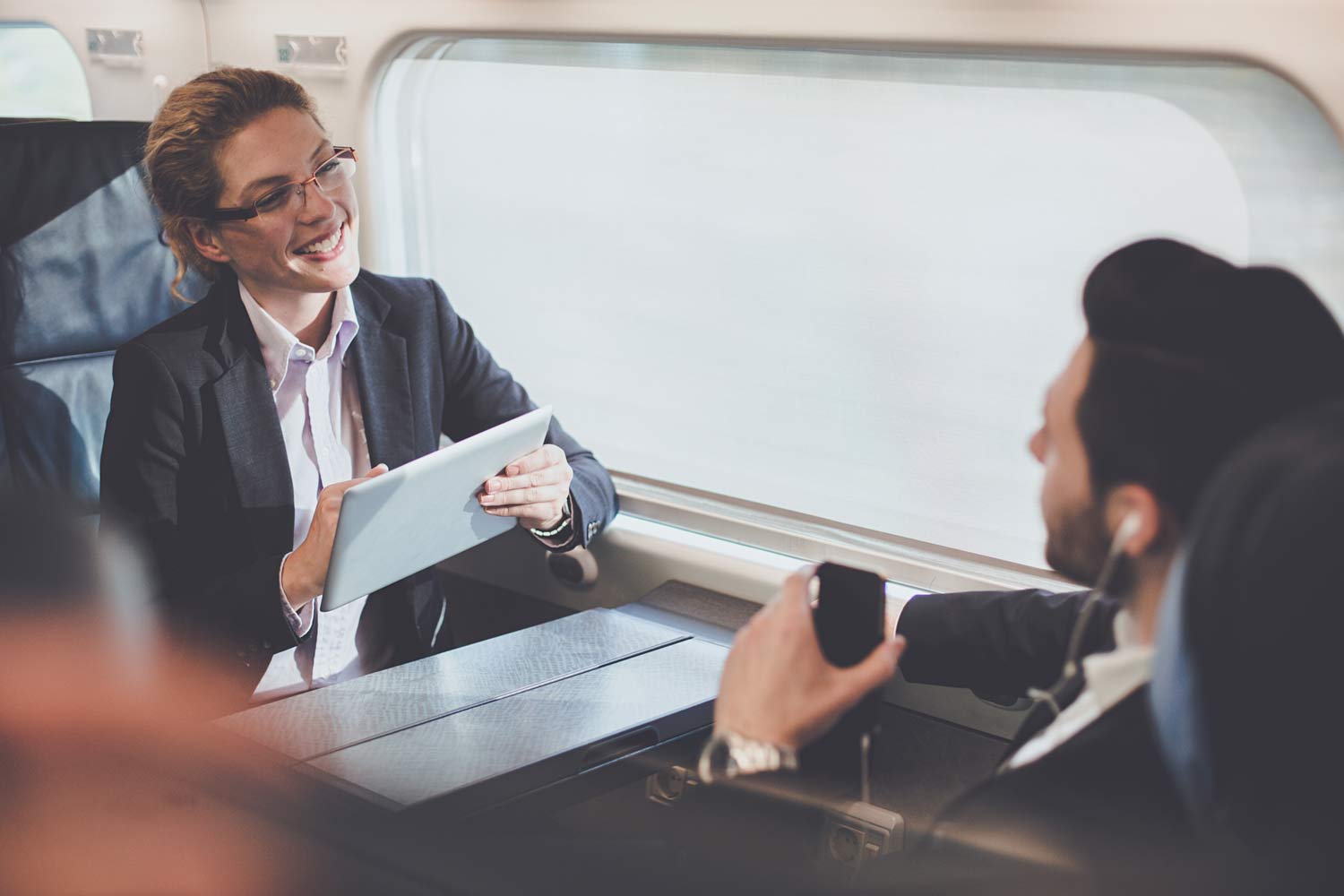 Mulher executiva com tablete e homem ao telemóvel, frente a frente numa mesa de reuniões, junto à janela, no Alfa Pendular.