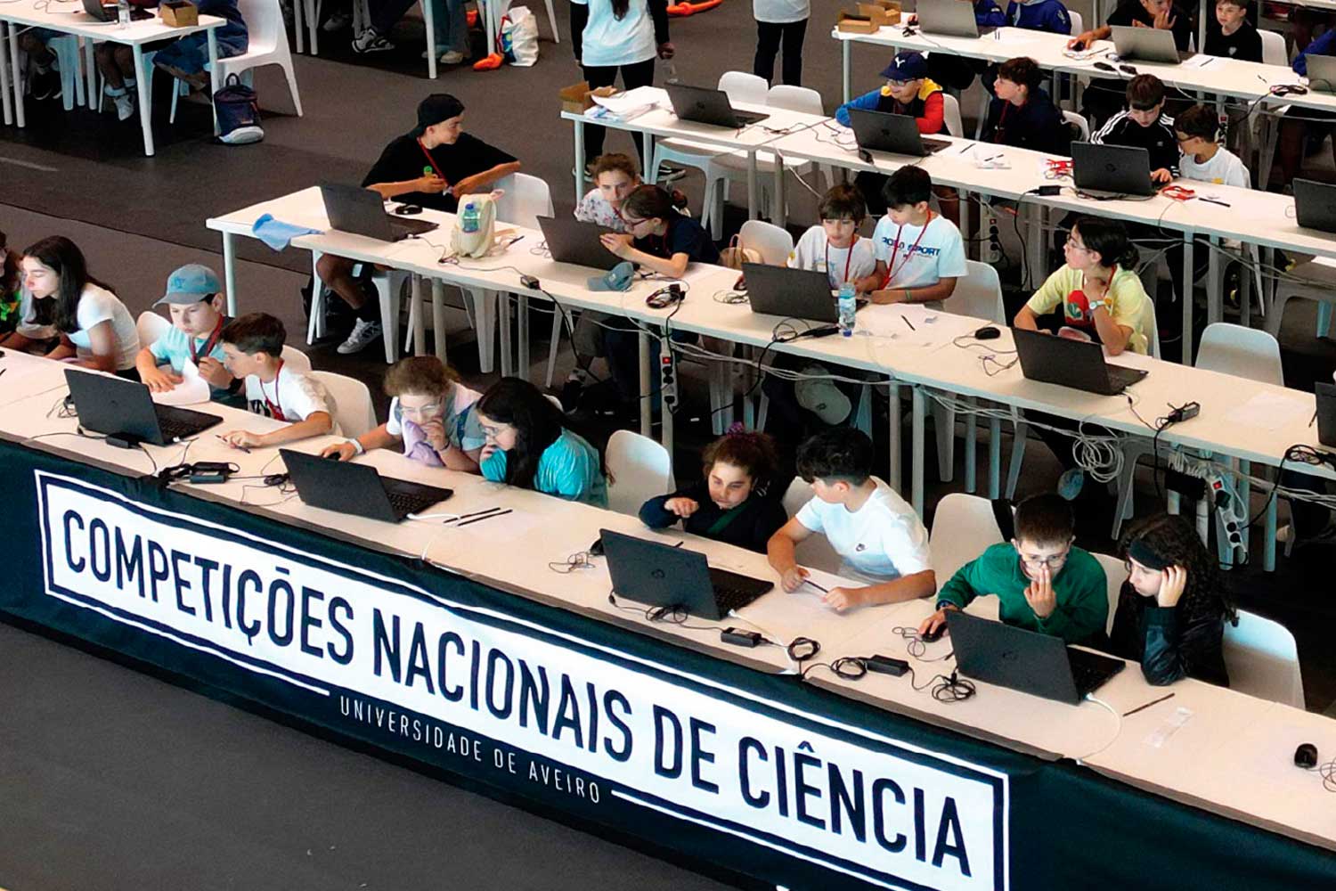 Alunos sentados em ambiente escolar e a trabalhar com computadores.