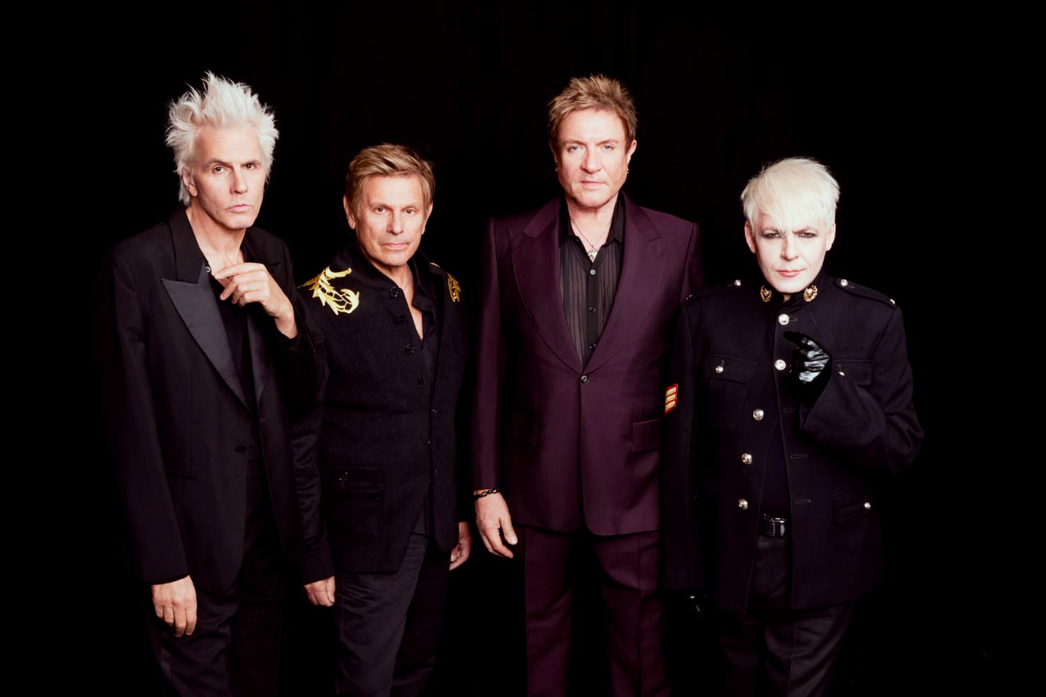 Imagem promocional dos Duran Duran, com os quatro membros da banda alinhados diante de um fundo preto, usando vestuário escuro e elegante.