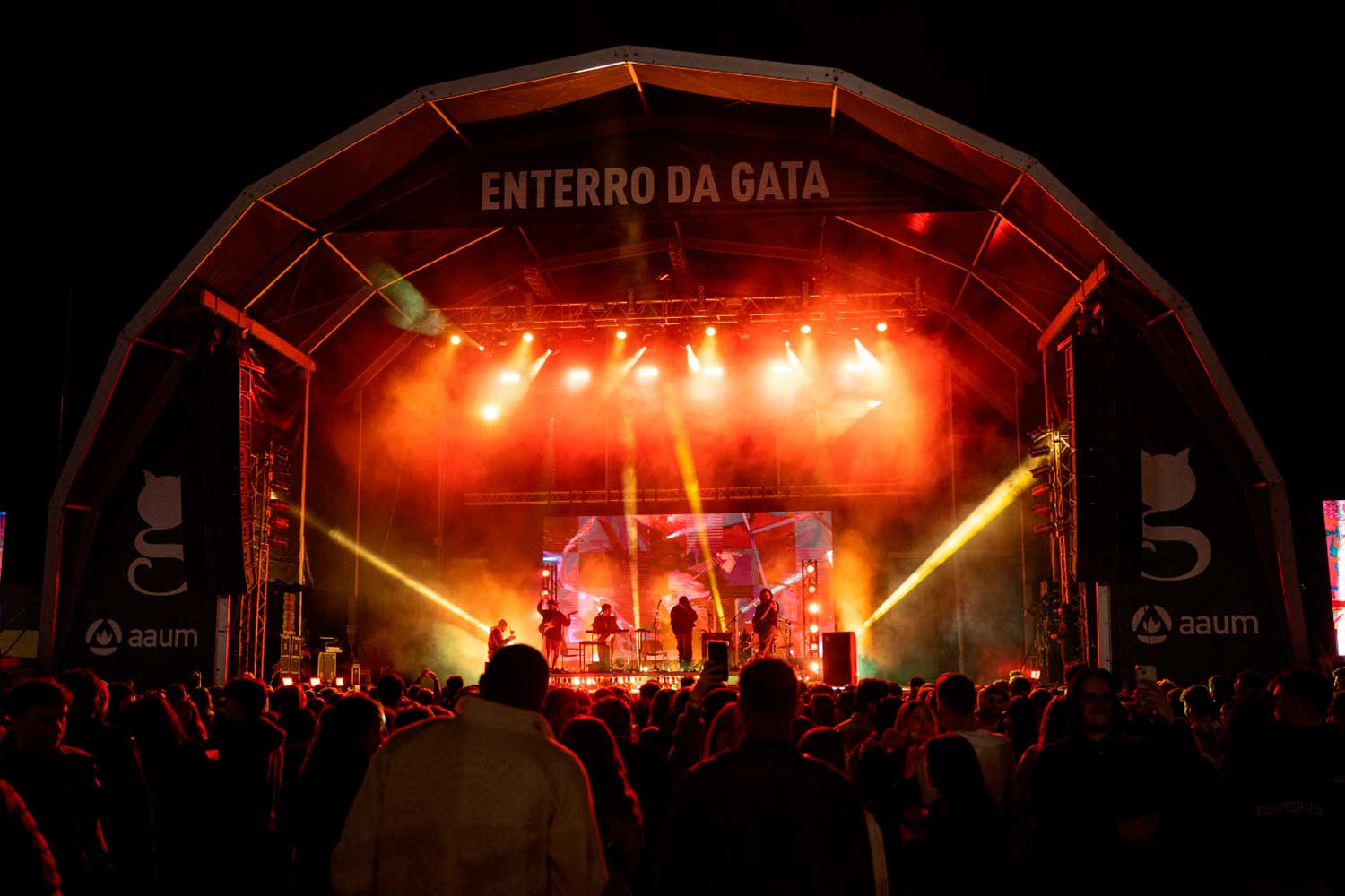 Imagem noturna de pessoas junto a palco, com banda a atuar.