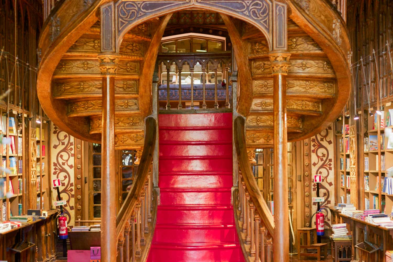 Imagem a cores de interior de livraria, com escada em madeira de acesso a piso superior e livros organizados em prateleiras de madeira.