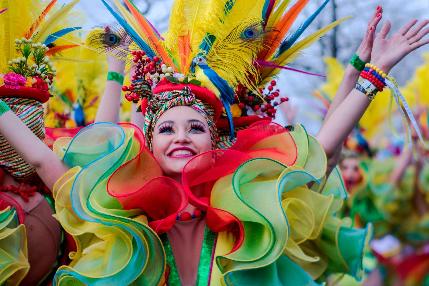 Participante de desfile com traje colorido e adereço de penas multicoloridas, rodeado por um ambiente festivo repleto de cor, criatividade e animação.